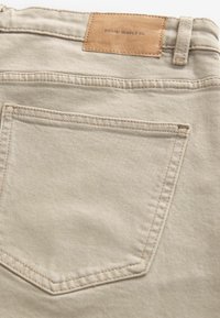 Beige denim jeans with a smooth texture, featuring a leather brand patch at the waistband and a single back pocket with contrasting stitching.