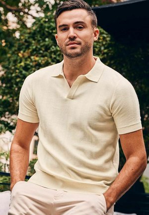 Young man in cream short-sleeve knit polo shirt and light pants, seated outdoors with greenery in the background.
