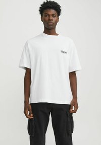 T-shirt bianco in cotone con vestibilità comoda, caratterizzato da una piccola scritta nera "original" sul lato sinistro del petto. Indossato con pantaloni cargo neri.