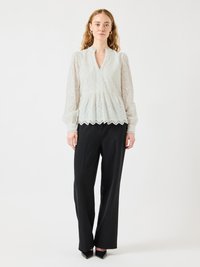 Blusa bianca in broderie anglaise con scollo a V, maniche a sbuffo e orlo scallop, abbinata a pantaloni neri a gamba larga e décolleté a punta.