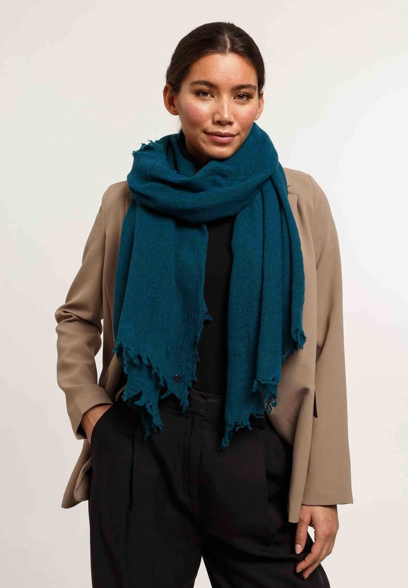Écharpe texturée teal avec des bords effilochés, drapée autour des épaules, associée à un blazer beige et un haut noir, offrant un look superposé.