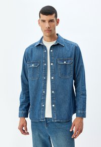 Camisa de mezclilla para hombre, de color azul oscuro, con dos bolsillos frontales, botones a presión y cuello extendido. Mangas largas con puños abotonados, de tejido de peso medio.