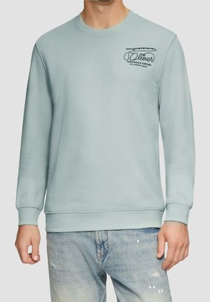 Mężczyzna w jasnoniebieskiej bluzie z okrągłym dekoltem z napisem "Oliver Hardware & Supplies Fly Fishing Equip" i grafiką na klatce piersiowej, ubrany w wyblakłe dżinsy.