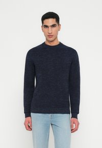 Man wearing a dark blue knit sweater and light blue jeans standing against a plain white background.
