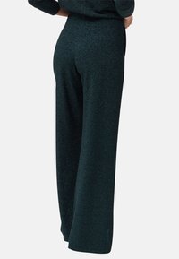 Pantalon vert foncé taille haute à jambes larges porté par une personne vue de dos, les mains sur les hanches, sur fond blanc.