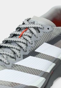 Γκρι αθλητικό παπούτσι με υφή στο σχέδιο, διαθέτει τρεις λευκές ρίγες, γκρι κορδόνια και πορτοκαλί θηλιά κορδονιού ως διακριτική λεπτομέρεια.