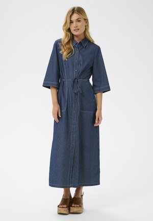 Long, blue denim dress with white stripes, collar, short wide sleeves, button front, waist tie, and two front pockets. Wood platform shoes.