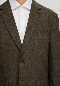 Brauner Tweed-Blazer mit Reverskragen, einem Knopfverschluss und aufgesetzten Taschen. Darunter ein weißes Hemd aus strukturiertem Stoff.