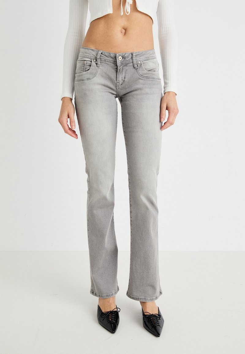 Femme portant un jean gris clair droit, un top blanc court à manches longues et des chaussures noires pointues à lacets, debout devant un fond uni.