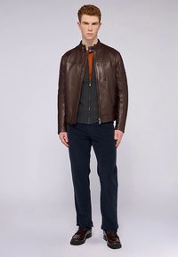 Chaqueta de cuero marrón con cuello alto, cierre frontal con cremallera y bolsillo lateral, sobre una camisa gris con un toque naranja, combinada con pantalones oscuros.