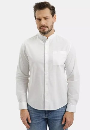 Mężczyzna z brodą, ubrany w białą koszulę zapinaną na guziki z kołnierzem typu mandarynka i kieszenią na piersi oraz niebieskie dżinsy, stoi na jednolitym tle.