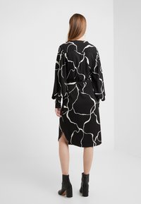 Schwarzes Kleid mit einem weißen abstrakten Muster, langen Ärmeln und einem taillierten Bund. Mit einem Schlitz im Rücken. Kombiniert mit schwarzen Knöchelstiefeln.