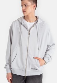 Jasnoszara bluza z kapturem zapinana na zamek, o gładkiej teksturze, z kieszeniami typu kangur oraz sznurkiem w kapturze. Z przodu znajduje się detal z logo.