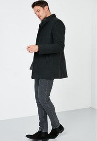 Abrigo negro de mezcla de lana con cuello alto y botones, combinado con jeans ajustados grises y botines de ante negros, sobre una modelo.