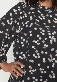 Schwarzes Stoffoberteil mit weißem Gänseblümchenmuster, das einen hohen Kragen und puffige Ärmel aufweist und einen strukturierten, leichten Look hat.