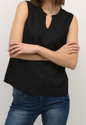 Femme portant un haut noir sans manches avec une encolure en encoche et un jean bleu, bras croisés à la taille sur un fond uni.