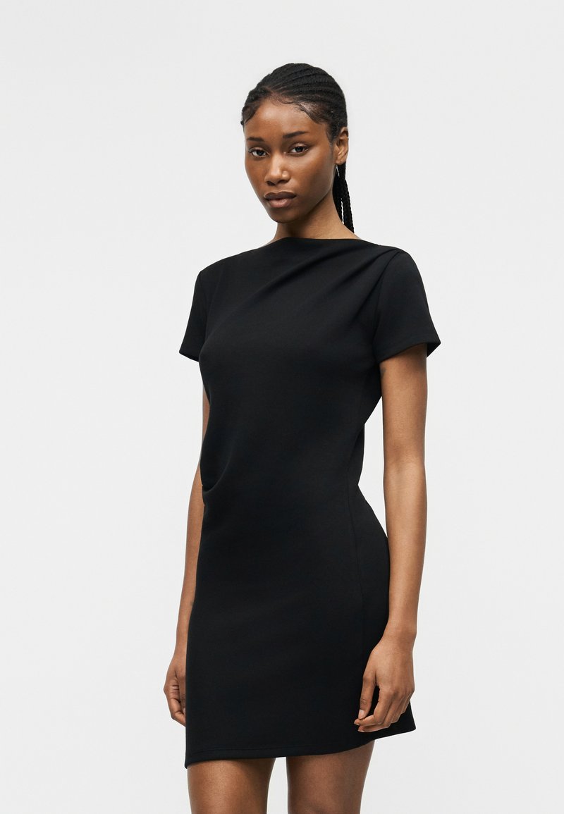 Γυναίκα με πλεξούδες, που φορά κοντό μαύρο εφαρμοστό φόρεμα με κοντά μανίκια, στέκεται σε απλό ανοιχτό φόντο.