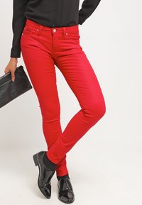 Czerwone wąskie dżinsy wykonane z gładkiego materiału o gładkim wykończeniu. Posiadają kieszeń zapinaną na zamek, pięć kieszeni oraz zapięcie na guzik. Połączone z czarnymi butami.