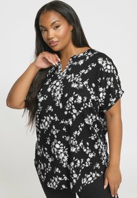 Kvinna med långt mörkt hår som bär en svart t-shirt med kort ärm och vit blommönster, poserar med en hand nära ansiktet.