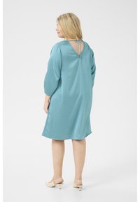 Robe teal avec une texture lisse, manches 3/4 et un dos en scoop avec une fermeture à nouer. L'ourlet est légèrement incurvé et tombe au-dessus du genou.