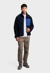 Fleecjacke in Marineblau mit blauen Akzenten und einer Reißverschlusstasche auf der Brust; getragen über einem weißen Hemd, kombiniert mit khakifarbenen Hosen und grauen Sneakern.