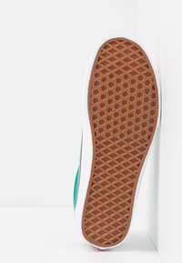 Sula av en avslappnad sneakers med brun våffelmönstrad sula, vit mellansula och teal-övre, placerad vertikalt mot ett vitt hörn.