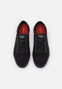 Zapatillas de lona negras con punta redonda, diseño con cordones y suela de goma. El interior presenta una plantilla roja con la marca. Los acentos incluyen un logo de bandera de colores.