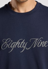 Camiseta de algodón azul marino con cuello redondo; presenta texto bordado en plata "Eighty Nine" en la parte frontal, textura suave y corte estándar.