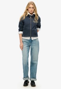 Sudadera navy con cremallera y cordón, logo dorado en el pecho, llevada sobre una camiseta blanca. Combinada con jeans de un azul claro y zapatos negros.