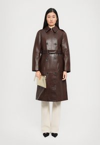 Donna che indossa un trench coat in pelle marrone scuro con cintura, tiene in mano una clutch beige, abbinato a pantaloni crema a gamba larga e scarpe nere a punta.