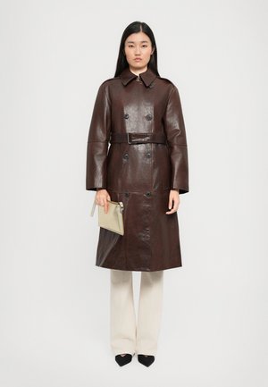 Donna che indossa un trench coat in pelle marrone scuro con cintura, tiene in mano una clutch beige, abbinato a pantaloni crema a gamba larga e scarpe nere a punta.
