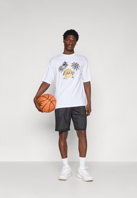 Biela bavlnená tričko s grafikou "Los Angeles Lakers" a palmami, skombinované s čiernymi sieťovými šortkami. Krátke rukávy, voľný strih a biele tenisky.