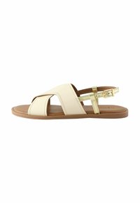 Sandal beige con design a incrocio, sottile cinturino alla caviglia, suola marrone chiaro e accentuazione dorata metallica sul cinturino. Texture del materiale liscia.