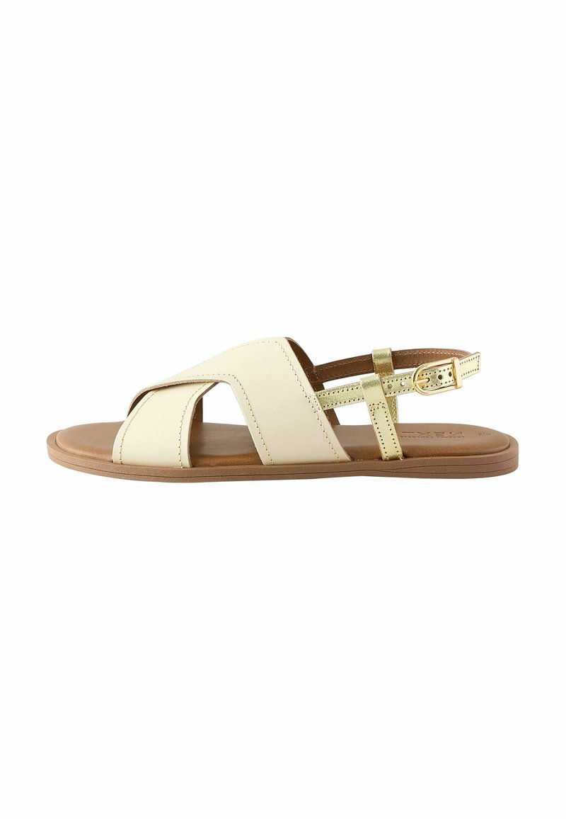 Sandal beige con design a incrocio, sottile cinturino alla caviglia, suola marrone chiaro e accentuazione dorata metallica sul cinturino. Texture del materiale liscia.