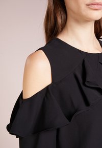 Femme portant un haut noir à épaules dénudées avec un détail à volants, montrant l'épaule nue ainsi qu'une partie du cou et du menton, sur un fond uni.