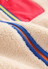 Kremowy polar z akcentami w kolorze czerwonym, niebieskim i pomarańczowym, o fakturowanej powierzchni i kontrastujących zamkach błyskawicznych.