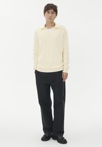 Világos sárga kötött pulóver V-nyakkal és gallérral, fekete, laza fazonú nadrággal és fekete fűzős cipővel párosítva. Egyszerű dizájn, puha textura.