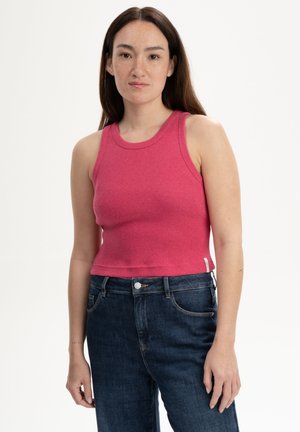 Vrouw met lang bruin haar, gekleed in een mouwloze roze top en donkerblauwe jeans, staand tegen een witte achtergrond.