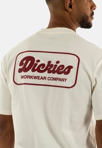 Kremo spalvos medvilnės marškinėliai su dideliu raudonu logotipu ant nugaros, kuriame parašyta "Dickies WORKWEAR COMPANY", trumpomis rankovėmis ir laisvu sijonu.