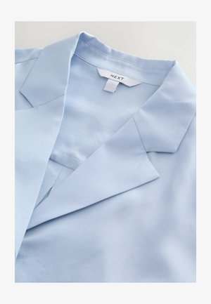 Tessuto azzurro chiaro, liscio, con collo con intagli e rialzo del colletto. Il capo presenta una leggera lucentezza, che mette in risalto il design strutturato ma rilassato.