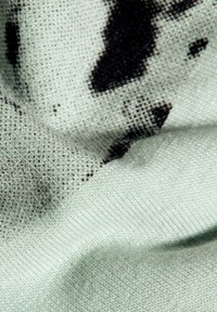 Grön tyg med en fin struktur, prytt med ett svart sprutmönster. Materialet verkar mjukt och något töjbart. Närbild.