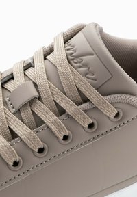 Sneaker beige avec une tige en synthétique lisse, bout rond, lacets texturés et un patch logo. Les coutures et les œillets sont subtilement visibles.