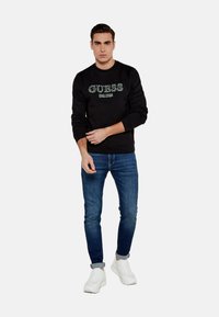 Felpa nera con logo "GUESS" in verde, abbinata a jeans blu scuro e sneakers bianche, caratterizzata da una vestibilità slim e scollo a girocollo.