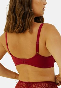 Soutien-gorge bordeaux avec bretelles réglables, finition en tissu lisse et fermeture à crochets. Détails en dentelle visibles le long de la bande.