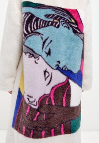 Fauxfurjacka i krämfärg med ett färgglatt abstrakt mönster på ryggen i rosa, blå och bruna nyanser, som föreställer stiliserade ansikten och djur.