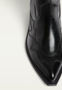 Stivale alla caviglia in pelle nera con punta affusolata, caratterizzato da cuciture testurizzate e motivi curvi lungo i lati. Finitura liscia e suola resistente.