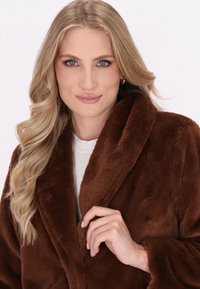 Donna bionda con occhi azzurri, che indossa un cappotto in faux fur marrone, tiene il colletto con la mano destra, sfondo neutro.