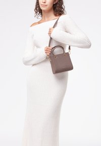 Borsa a tracolla in cuoio marrone con texture, hardware dorato, manici corti e tracolla staccabile, portata da una persona in un vestito bianco.
