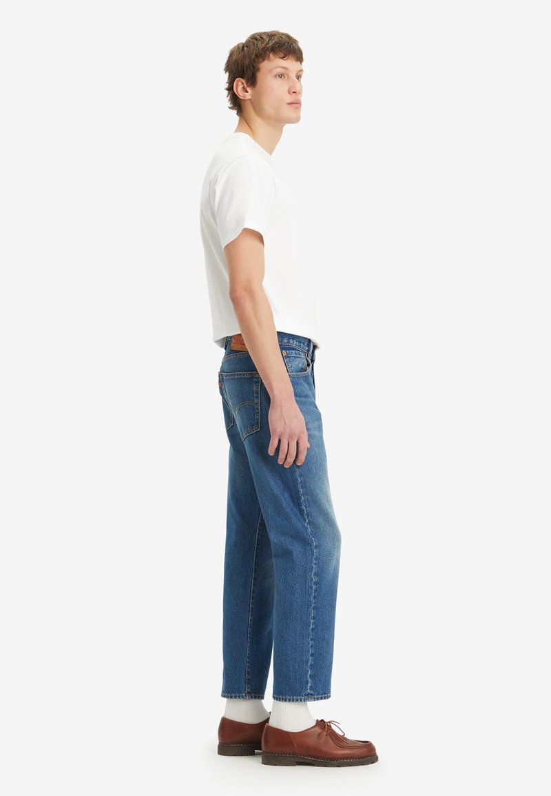 Camiseta blanca de manga corta, jeans de mezclilla azul de corte holgado y textura desgastada, zapatos de laceado marrón y calcetines blancos, vista de perfil.