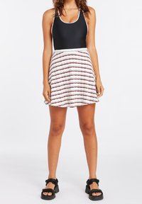 Top in costume da bagno senza maniche nero abbinato a una gonna bianca con strisce ondulate rosse e blu. Le calzature consistono in sandali neri con zeppa.
